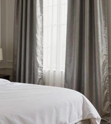 A professional interior photography shot of an upscale South Asian / Indian living space featuring premium home textiles. High-thread-count white bedsheets and elegant silver-toned curtains are showcased with soft, natural lighting creating an atmosphere of sophistication.