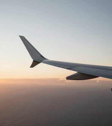 An artistic, high-contrast shot of an airplane wing slicing through a sunset sky over the Atlantic. Golden hour light hitting the wing, minimalist and aspirational, #FAF8F5 and #2C3539.