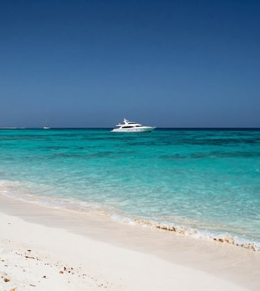 Pristine turquoise waters of a Red Sea resort in Egypt. Alabaster white sand in the foreground and a luxury yacht floating in the distance under a clear deep blue sky.