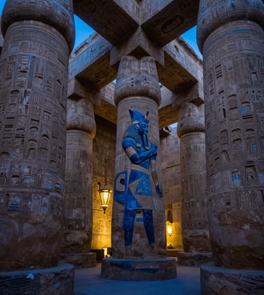 The interior of Luxor Temple at dusk, showing the massive sandstone columns. Deep Egyptian blue shadows fill the spaces between pillars, while gilded gold lantern light illuminates the detailed hieroglyphs.