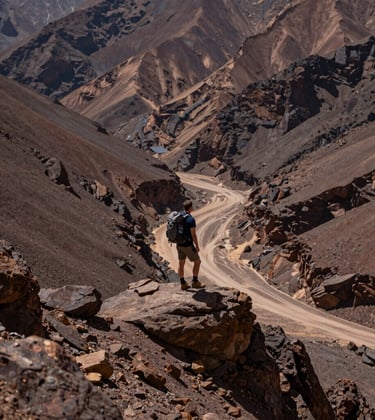 A hiker standing on a ridge in the Atlas Mountains, looking out over a valley with deep espresso colored rocks and soft sand paths.