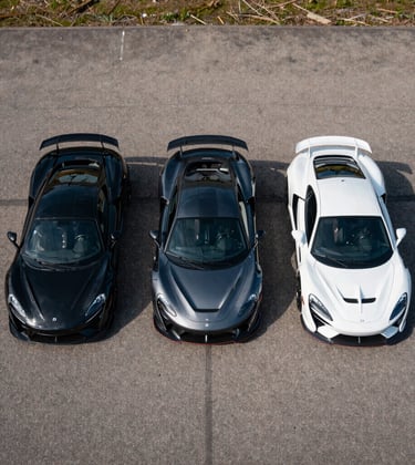 An aerial view of three high-performance sports cars in obsidian black, deep slate charcoal, and pristine cool grey parked in perfect alignment on a private tarmac.