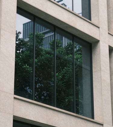 Architectural detail of a modern financial center building. Clean lines, soft off-white stone surfaces, and large glass panels reflecting a forest green plant nearby.