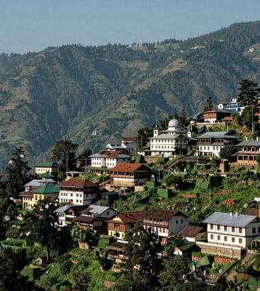 A breathtaking view of the mountain town of Shimla, featuring colonial-style architecture nestled among lush green ridges under a clear sky. The composition captures the serene beauty of the South Asian highlands in bright, natural daylight.