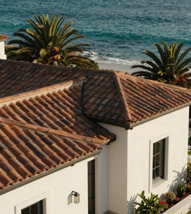 A Mediterranean-style luxury villa in La Jolla featuring muted bronze tiled roofs and off-white stucco walls, set against a backdrop of lush green palms and turquoise water.