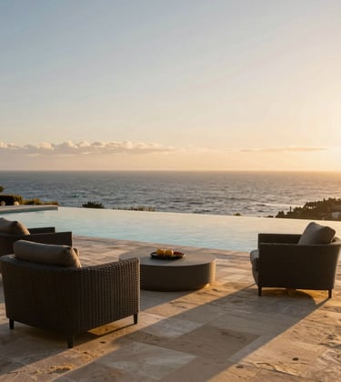 An infinity pool overlooking the Pacific Ocean at sunset, with Deep Charcoal outdoor furniture and Warm Sand stone tiling. The vibe is aspirational, exclusive, and peaceful.