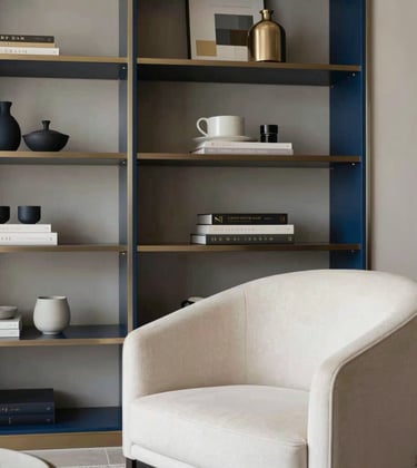 A detailed shot of a luxury interior solution featuring a custom-built shelving unit and a minimalist armchair in a soft off-white fabric. The setting is a sophisticated South Asian / Indian apartment with subtle steel blue accents and artistic decor, capturing an atmosphere of refined elegance and modern living.