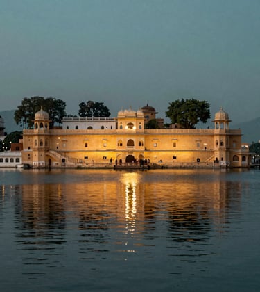 A vibrant photography shot of the City Palace in Udaipur reflecting in the still waters of Lake Pichola at dusk, showcasing South Asian / Indian heritage with elegant lights and a muted teal sky.