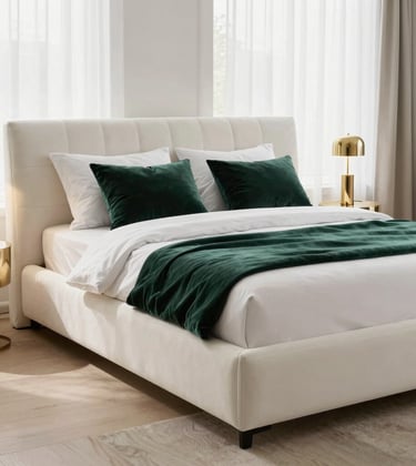 A luxury master bedroom featuring a plush soft white upholstered bed, dark green velvet throw pillows, and metallic gold bedside lamps. The room is airy with sheer white curtains and beige hardwood flooring.