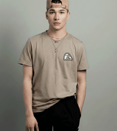 Joven usando una camiseta color beige de cuello redondo y una gorra, posando frente a un fondo gris.