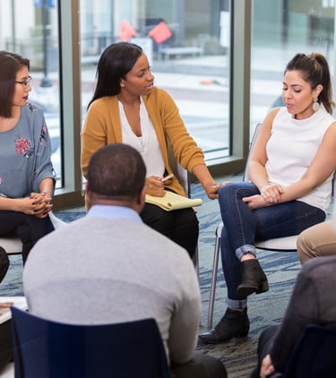 a group of people sitting in a circle with a woman in a white shirt