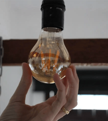 Hand holding a glowing vintage Edison light bulb hanging from a rustic wooden beam fixture.
