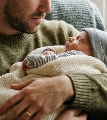 Father hold newborn baby in his arms. Father wears and wedding ring and sits in a UK living room.