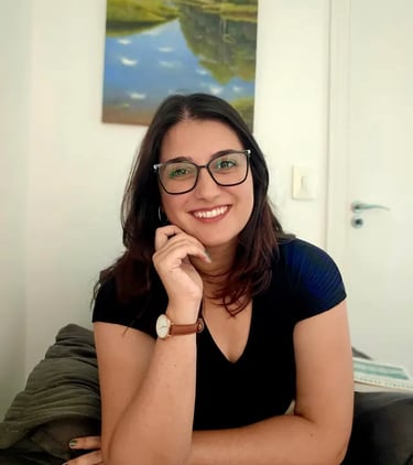 Psicóloga Rafaela Antoniazzi sorrindo, vestindo óculos e blusa preta, apoiando o rosto na mão. Seção Sobre.
