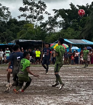 Athletes playing a competitive soccer match on a muddy rural field surrounded by spectators.