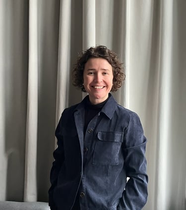 Smiling woman with curly hair wearing a blue jacket standing against light grey curtains.