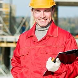 Smiling male construction worker in a red uniform and yellow hard hat holding a clipboard.