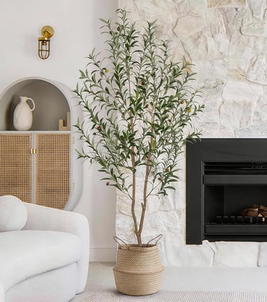 Tall artificial olive tree in a woven basket planter standing in a modern living room next to a fireplace.