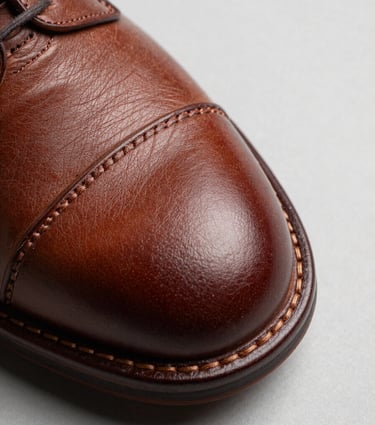 Close-up macro of a leather shoe sole edge and upper stitching seam on a neutral light grey surface, studio strobe light raking across the leather grain to reveal texture and precision, overhead slight angle, no models, no branding
