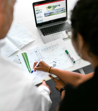 Two architects reviewing landscaping blueprints and site plans on a desk with a laptop.