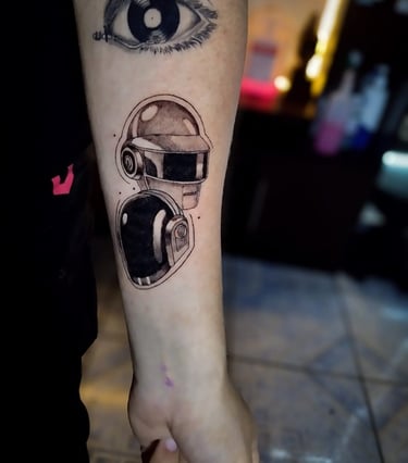 Tatuaje de los cascos de Daft Punk en un antebrazo, debajo de un tatuaje de un ojo surrealista.