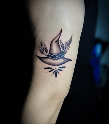 Tatuaje de sombrero de bruja en tinta negra en el brazo de una persona, con hojas botánicas.