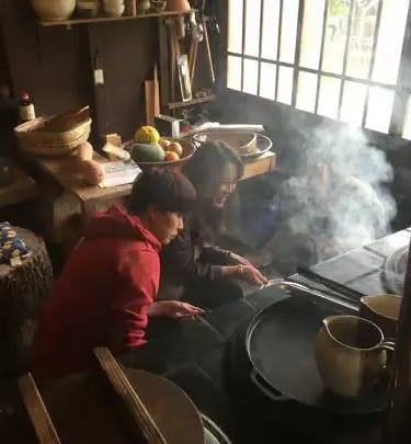 Two people cooking over an open hearth in a rustic Japanese farmhouse kitchen