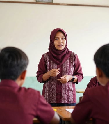 Candid eye-level portrait of a local Indonesian teacher explaining a concept to an engaged student in a bright, clean classroom, warm daylight, rich maroon details, 35mm.