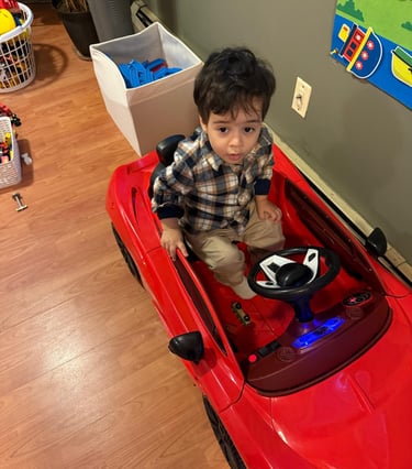 A young boy sitting in a red electric ride-on toy car in a playroom.