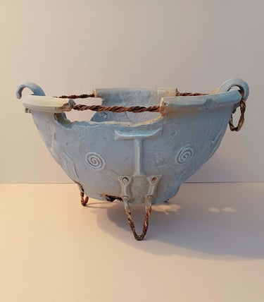 A fragmented, round bowl-like sculpture adorned with symbols and letters, finished in blue-grey hues