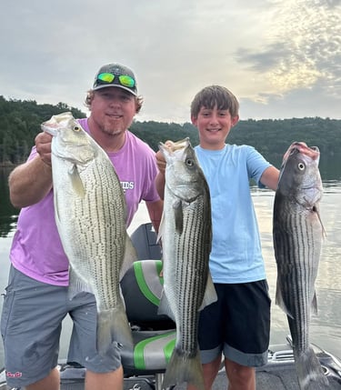 A man and boy smiling on a boat while holding up three large striped bass caught during a lake fishing trip.