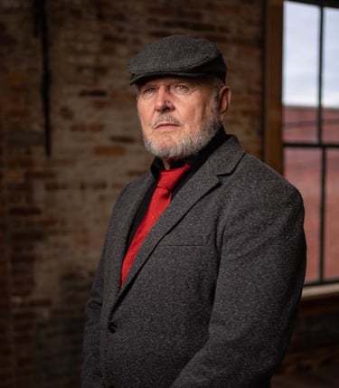 Portrait of Houston Baker wearing a grey wool blazer, flat cap, and red tie in a rustic loft.