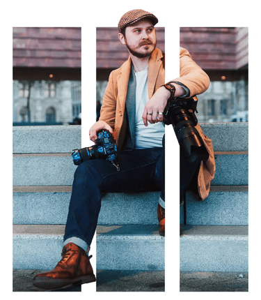 Professional photographer in a flat cap and tan coat holding a DSLR camera on city steps.