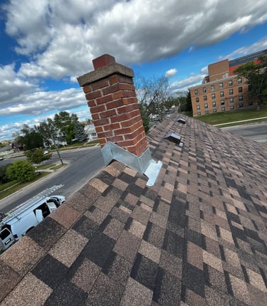 Gabled roof peak, brown asphalt shingles and chimney flashing. Roof vents and brick chimney. 
