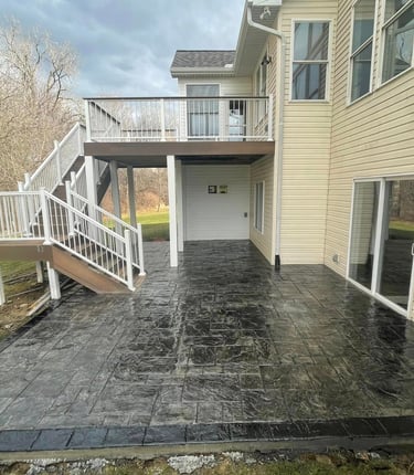New dark grey stamped concrete patio installed under a second-story wooden deck with white railings.