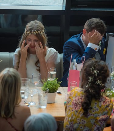 An emotional bride and groom crying during wedding speeches at a rustic reception table.