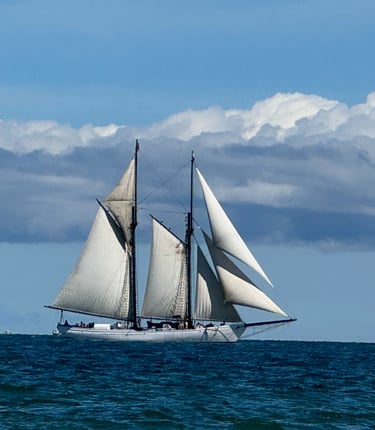 A classic two-masted schooner sailing on blue ocean water under a cloudy sky.