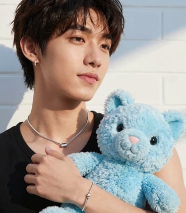A close-up shot of a chic young individual from the North American / Global Trendy Demographic holding a cute pastel blue plush toy and wearing trendy minimalist silver jewelry, standing in front of a clean white brick wall with soft sky blue shadows. High fashion, warm natural sunlight, joyful and playful charm.
