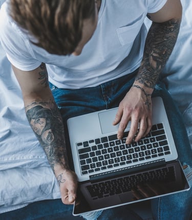 man with tattoos on arms holding a laptop while sitting on a bed