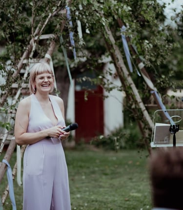 Traurednerin Silke lacht während einer freien Trauung und hält eine persönliche Rede mit Mikrofon im Garten.