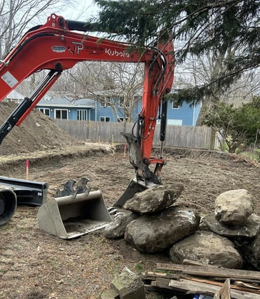 Orange Kubota excavator moving large boulders for a backyard landscape retaining wall project.