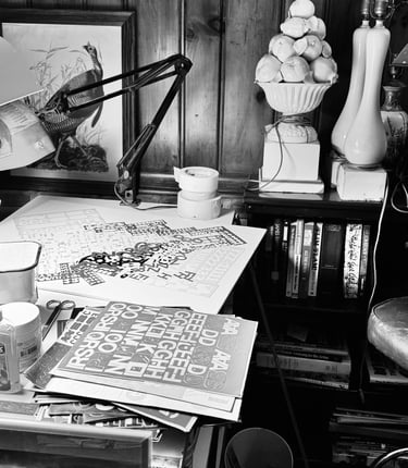 Cluttered artist's workspace with vintage desk supplies, art prints, and a leather chair in black and white.