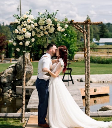 Newlywed couple kissing under a rustic wooden wedding arch decorated with white roses at an outdoor ceremony.
