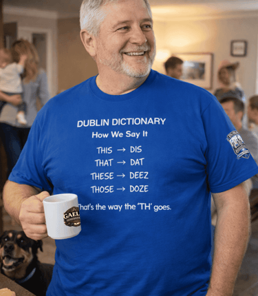 A smiling man wearing a blue Dublin Dictionary t-shirt with Irish slang pronunciation, holding a coffee mug.