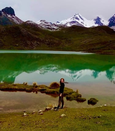 A hiker stands by a turquoise lake reflecting snow-capped peaks in the Andes mountains.