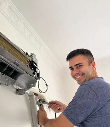 A smiling technician uses an adjustable wrench to repair an indoor wall-mounted air conditioning unit.