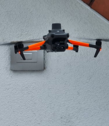 A DJI Mavic drone with orange arms flies near a building wall for an aerial roof inspection.