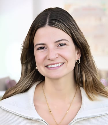 Smiling woman with long brown hair wearing a white zip-up sweater and gold jewelry in a bright room.