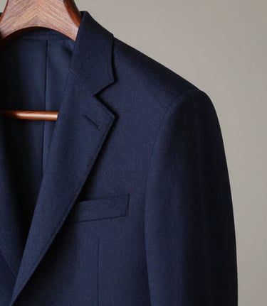 Chiaroscuro shot of a deep navy tailored suit jacket on a wooden hanger, soft side lighting highlighting structured shoulders and premium wool weave.