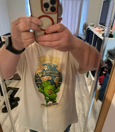 A man takes a mirror selfie wearing a graphic t-shirt featuring a green Irish mythological figure holding a globe.